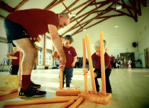 Two excited children help a didi cricket coach put up cricket stumps in a hall