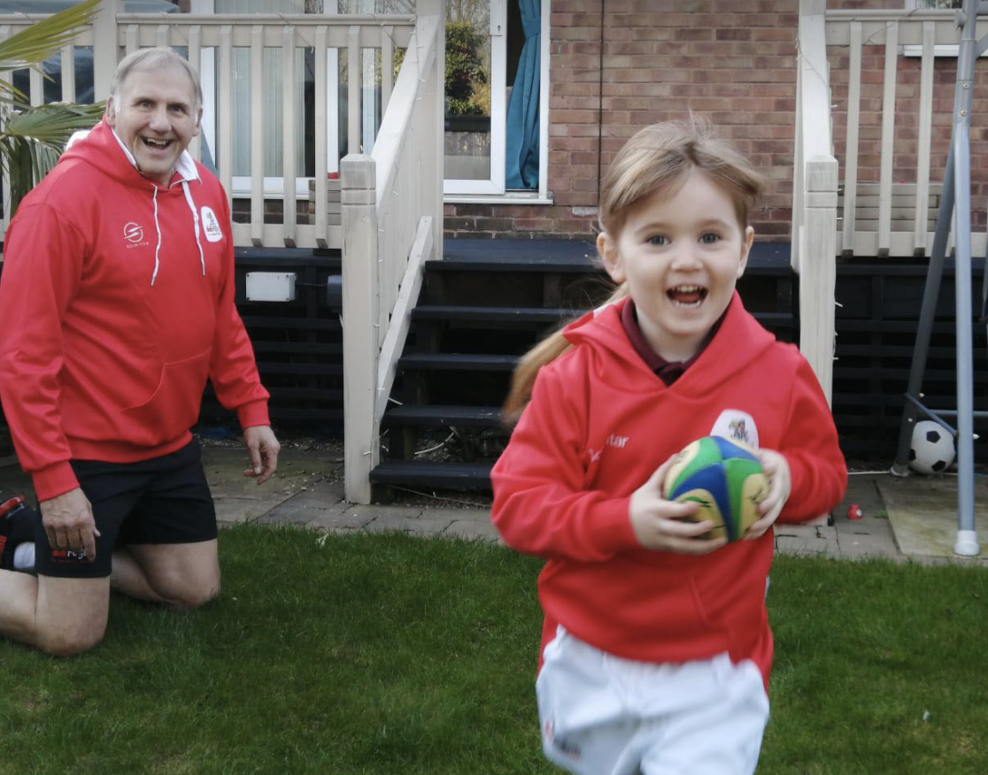 A small girl running with a rugby ball in her hand, smiling