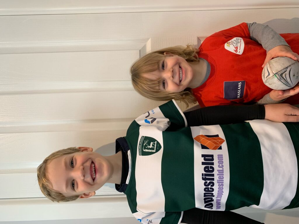 Two boys in rugby shirts smile with rugby balls in their hands