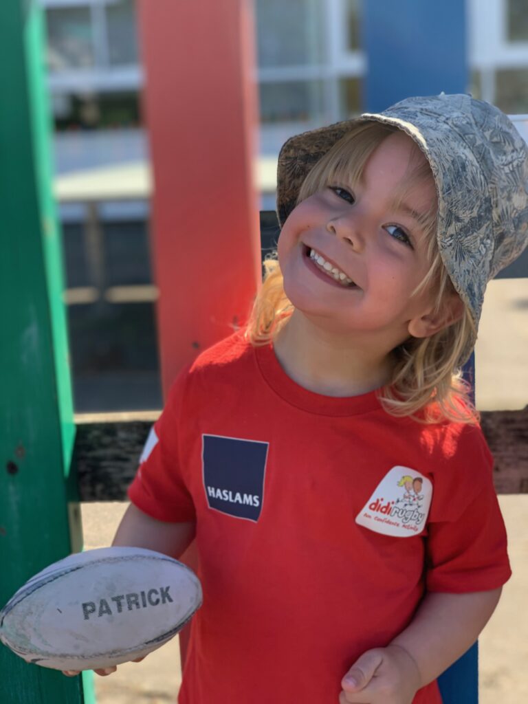 A you g boy in a red tee-shirt and sun hat smiles while holding a rugby ball 