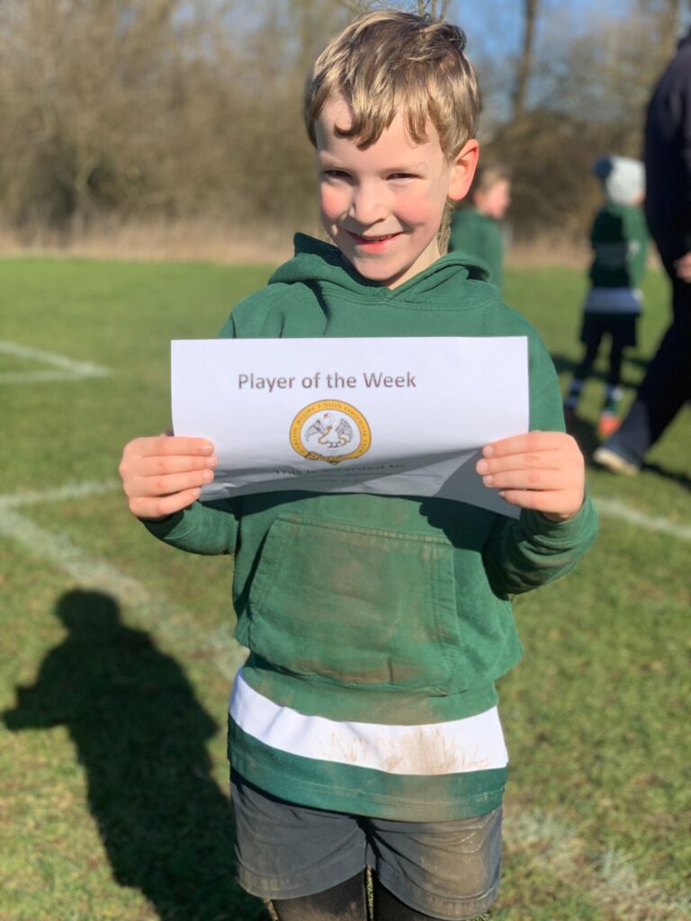 A young boy in a green hoodie holds a 'player of the week' certificate aloft