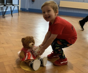 A boy in a red tee-shirt smiles as he picks up a rugby ball and teddy bear
