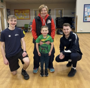 A boy in a green shirt is flanked by rugby coaches. Everyone is smiling