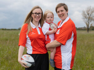 Sophie and Hattie Elliott-Edwards wearing red didi rugby kit, holding their son, Oliver.