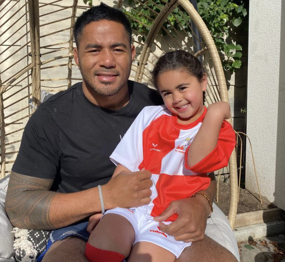 A man in a black shirt holds his young daughter who is wearing didi rugby kit