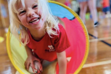 A blonde-haired girl crawls through a tunnel holding a rugby ball at one of our nurseries. She is smiling.