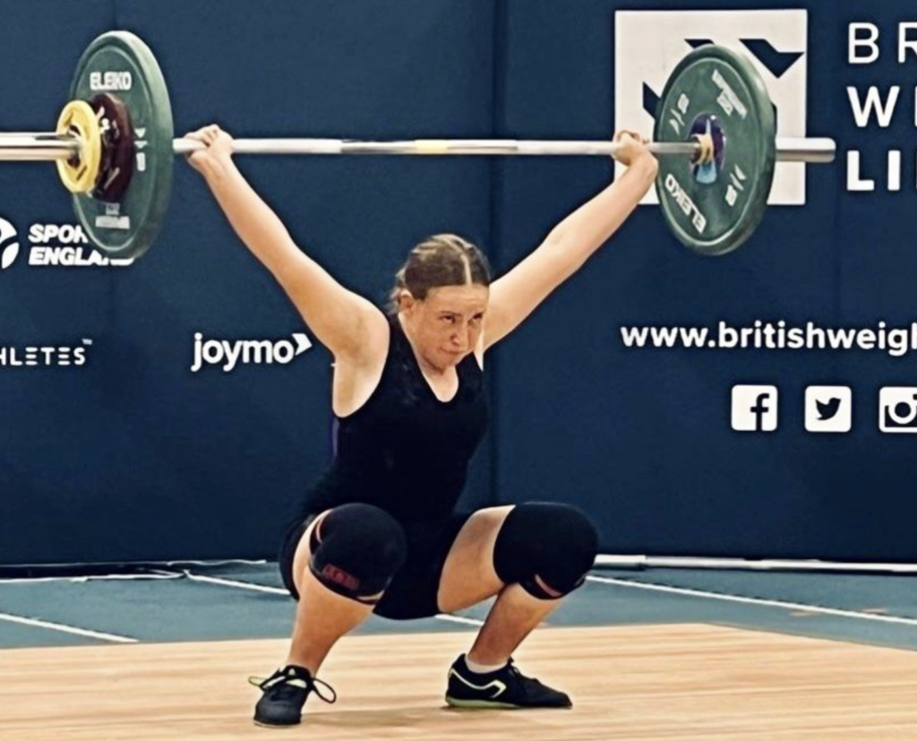 A woman in a blue leotard lifts a heavy weight above her head