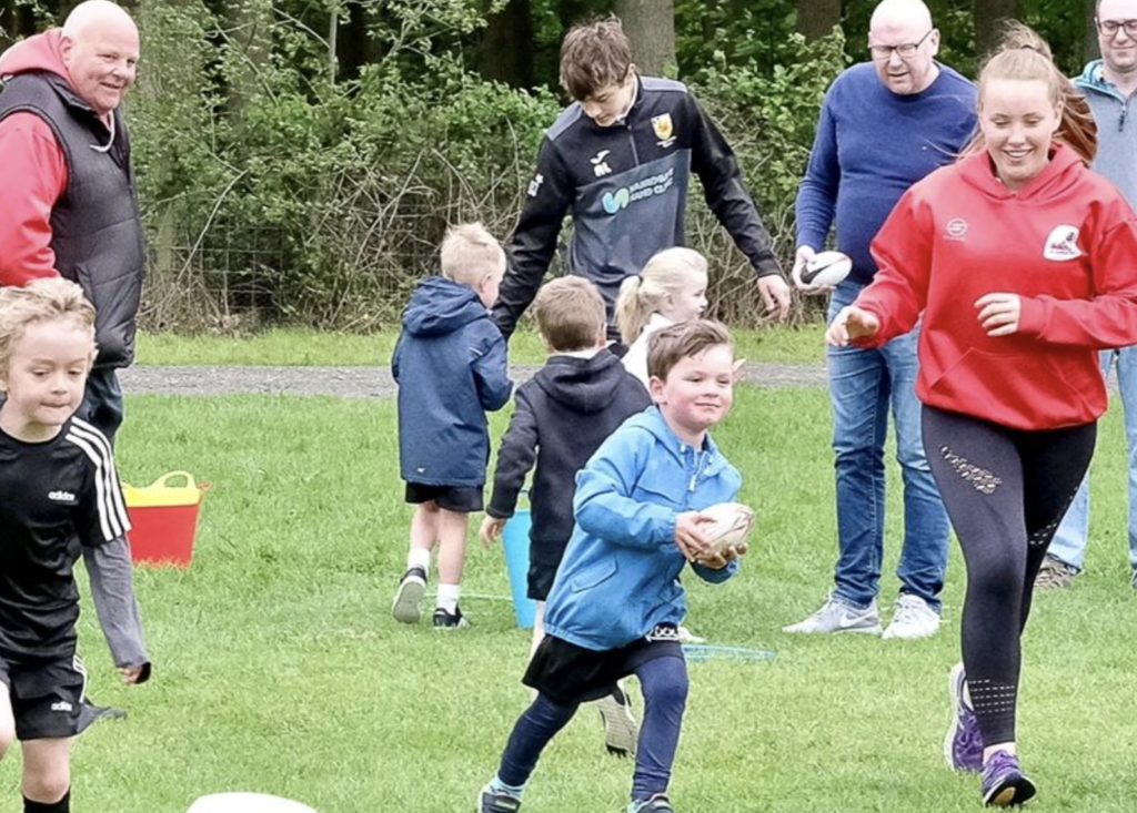 A lady in a red top runs alongside a young boy, smiling, holding a rugby ball